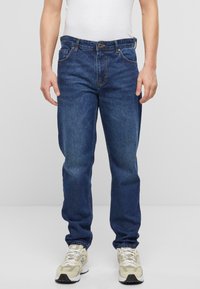 Jeans de mezclilla azul oscuro con corte recto, cinco bolsillos, una clásica cremallera con botones y detalles de costura visibles. Combinados con zapatillas beige.