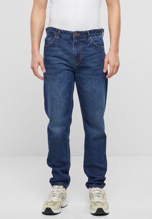 Jeans en denim bleu marine avec une coupe droite, cinq poches, une fermeture à bouton classique et des détails de couture visibles. Portés avec des baskets beiges.