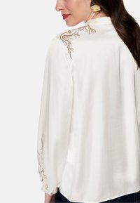 White satin blouse with long sleeves, featuring gold floral embroidery along the shoulders and sleeves, and a smooth, glossy texture.