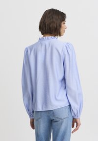 Lichtblauwe blouse met lange mouwen, een gestructureerde afwerking, ruches bij de hals en geplooide pofmouwen; gecombineerd met lichtblauwe jeans.