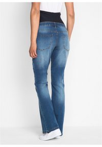 Jeans in denim blu con una vita scura, design a gamba svasata, leggero sbiadimento sulle cosce e due tasche posteriori visibili. Scarpe bianche sotto.