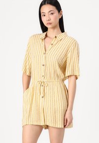 Robe courte à rayures jaunes avec des manches courtes, patte de boutonnage à l'avant, taille à cordon et poches latérales. Le tissu a une finition légère et texturée.