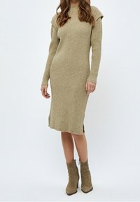 Robe tricotée couleur marron clair avec texture côtelée, manches longues, détails sur les épaules, fentes sur les côtés, associée à des bottines beige. 