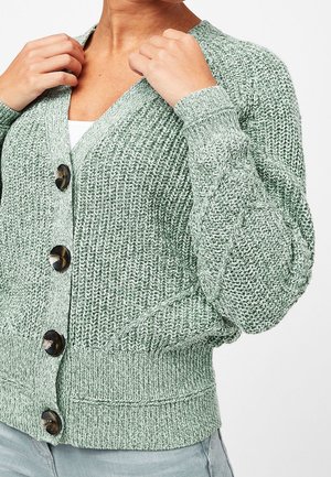 Femme ajustant un cardigan vert clair en maille épaisse avec de gros boutons foncés, poignets et ourlet côtelés, porté par-dessus un haut blanc et un pantalon gris clair.