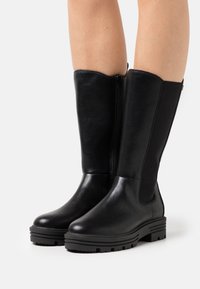 Bottes hautes jusqu'au genou en cuir noir avec une texture lisse, fermeture éclair latérale et panneau élastique contrastant. Semelle en caoutchouc épaisse avec adhérence.