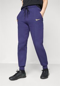 Lilla joggebukser laget av bomullsblanding, med elastisk midje med snor, sidelommer og en gul Nike-logo på venstre lår.