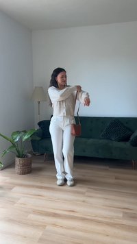 Cardigan de malha creme com detalhes em botão, calças brancas e pequena bolsa de ombro castanha. Sofá verde e candeeiro ao fundo, piso de madeira.