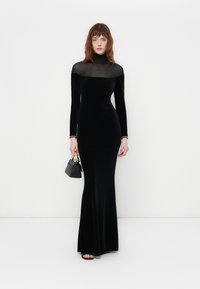 Black velvet long-sleeve dress with a sheer neckline and fitted silhouette. Paired with a small black handbag and open-toe sandals.