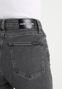 Vaqueros de mezclilla gris con un parche de cuero que incorpora el logotipo "CALVIN KLEIN JEANS", acentuados con un diseño de costura distintivo en el bolsillo trasero.