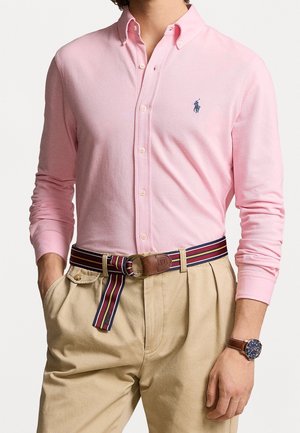 Man draagt lichtroze overhemd met knopen en marineblauw logo, beige broek, gestreepte stoffen riem met lederen detail en een bruine horloge met blauwe wijzerplaat.