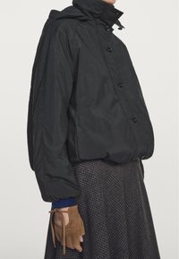 Zwarte gewatteerde jas met hoge kraag en knopen. Gecombineerd met een bruine geruite rok en vingerloze suède handschoenen.