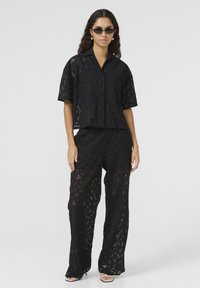 Ensemble en dentelle noire composé d'un haut à manches courtes avec col et d'un pantalon à jambe large, mettant en avant des motifs géométriques et une texture transparente.