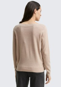 Vrouw met lang donker haar, gekleed in een beige lange mouw trui en donkere broek, van achteren weergegeven tegen een effen achtergrond.
