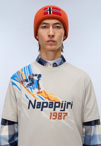 Beige t-shirt med blå og orange skiløbs grafik, tekst "Napapijri 1987"; lagd med en blåternet skjorte; rød strikket hue med norsk flag.