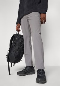 Pantalones grises de exterior con corte ajustado, acompañados de una mochila de tela negra. Se llevan zapatos deportivos negros con suelas texturizadas y cordones.