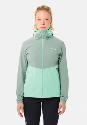 Vrouw draagt een tweekleurige groene capuchonjas met een ritssluiting aan de voorkant en ritssluitingen op de zijzakken, staand tegen een effen achtergrond.