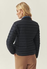 Veste noire matelassée avec motif matelassé horizontal, col montant et coupe ajustée. Portée par-dessus une chemise bleu clair à col.