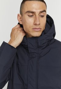 Mørkeblå pufferjakke med høy krage, quiltet tekstur og glidelåslommer. Modellen justerer kragen mens hun ser litt til siden.
