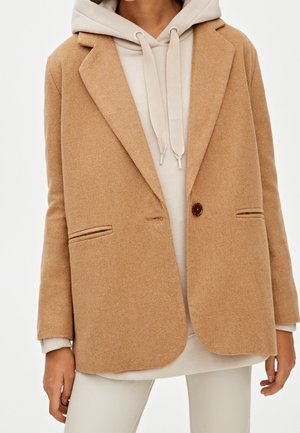 Femme portant un blazer en laine beige par-dessus un sweat à capuche beige clair et un pantalon assorti de couleur claire.