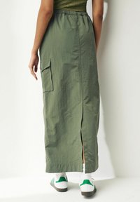 Person in einem langen olivgrünen Rock mit einer Seitentasche und einem Schlitz, kombiniert mit weißen Sneakern mit grünen Streifen und weißen Socken.