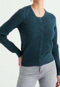 Teal knitted cardigan with a round neck, button front, textured pattern, and ribbed cuffs and hem, paired with light gray jeans.