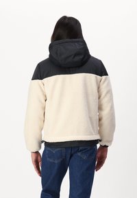 Fleece- ja nylontakki, jossa on kermaisen värinen vartalo, mustat olkapäät, säädettävä helma ja vuorillinen huppu joustavilla hihaosuilla.