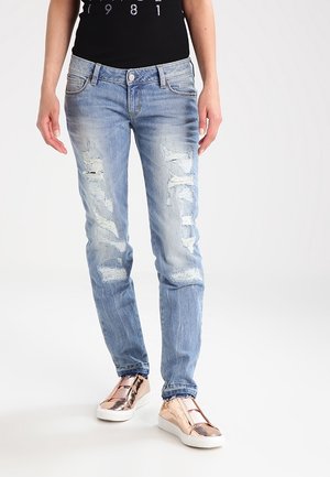 Persoon draagt lichtblauwe gescheurde spijkerbroek en metallic rozenkleurige slip-on sneakers tegen een witte achtergrond.