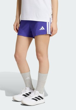 Beine einer Person, die lila Adidas-Shorts, graue Sportsocken und weiße Adidas-Sneaker mit schwarzen Streifen trägt und auf hellem Hintergrund steht.
