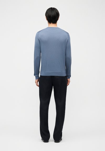 Άντρας που στέκεται με την πλάτη, φορώντας μπλε πουκάμισο με μακριά μανίκια και μαύρο παντελόνι, μπροστά σε απλό ανοιχτό φόντο.