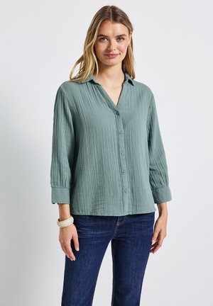 Donna che indossa una blusa button-up verde chiaro con texture e maniche 3/4, insieme a jeans blu scuro, in piedi davanti a uno sfondo bianco.