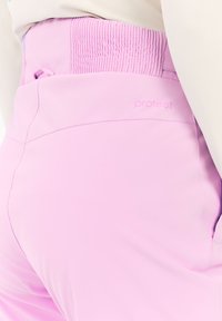 Close-up van een persoon die een lichtroze broek draagt met een hoge, elastische tailleband en een subtiele "protest" logo op de achterkant.