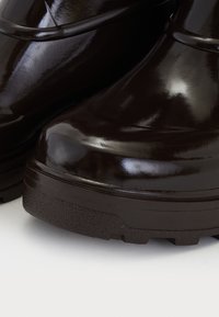 Botas de cuero negro brillante con suelas de goma gruesa y texturizada, primer plano en el área de los dedos contra un fondo neutro.