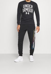 Felpa nera con il testo "UNDER ARMOUR" in bianco, abbinata a pantaloni della tuta neri con un logo multicolore sul lato sinistro. Sneakers bianche.