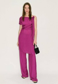 Magenta twee-delige outfit met een top met één schouder en gedrapeerde stof, gecombineerd met brede broekspijpen. Textuur zwarte handtas als accessoire.