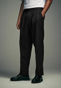 Black pinstriped trousers with a relaxed fit, featuring side pockets and a waistband. Worn with green loafers, showcasing a smooth texture.