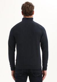 Tumesinine pullover-jakk kõrge kraega, soonilise tekstuuriga ja pikkade varrukatega. Omab lihtsat õmblust ning diskreetset logo õlal.