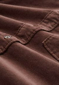 Bruine stof met een zachte, gestructureerde afwerking. Heeft zichtbare stiksels en een knoopdetail, wat een casual designesthetiek uitstraalt.
