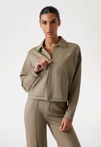 Khaki kurtka zapinana na zamek wykonana z gładkiego materiału, z kołnierzem, dwoma przednimi kieszeniami oraz wytłoczonym logo marki na piersi.