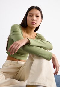 Crop top a maniche lunghe in maglia, di colore verde oliva, abbinato a pantaloni larghi di colore crema. Modella seduta, che mostra una postura rilassata.