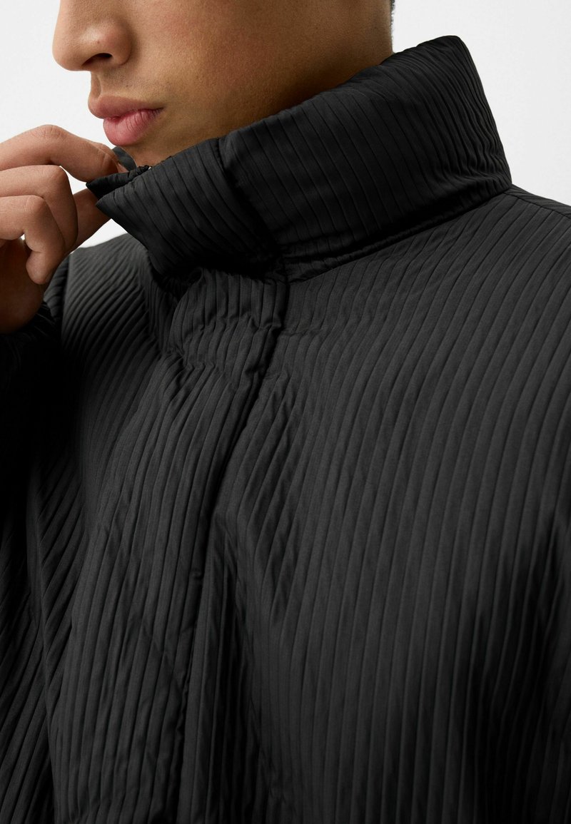 Schwarze, gerippte Jacke mit hohem Kragen. Das Material wirkt glatt mit sichtbarer Textur; verfügt über einen Reißverschlussdetails nahe dem Hals.