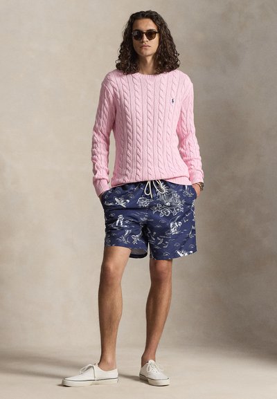 Man met lang krullend haar, een zonnebril, een roze kabelgebreide trui, marineblauwe korte broek met patroon en witte sneakers, staand met handen in de zakken.