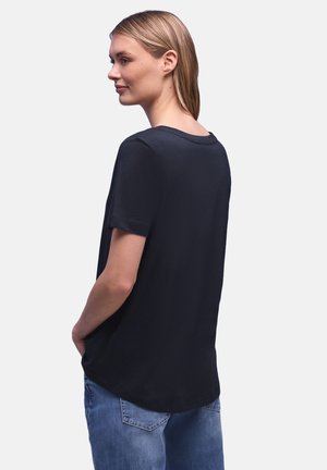 Jonge vrouw met recht blond haar, gekleed in een effen zwart T-shirt met korte mouwen en een blauwe spijkerbroek, staand met haar handen in haar zakken, licht naar links gedraaid.