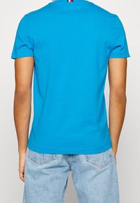 T-shirt di cotone blu con collo tondo, maniche corte e orlo dritto, caratterizzato da una piccola striscia rossa, bianca e blu sul retro del collo.