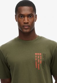 Camiseta de algodón verde oliva con mangas cortas. Presenta un texto gráfico rojo en el área izquierda del pecho. Cuello redondo con una textura suave.