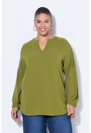 Lächelnde Frau mit kurzen Haaren, die goldene Creolen, eine olivgrüne Bluse mit langen Ärmeln und blaue Jeans vor einem weißen Hintergrund trägt.