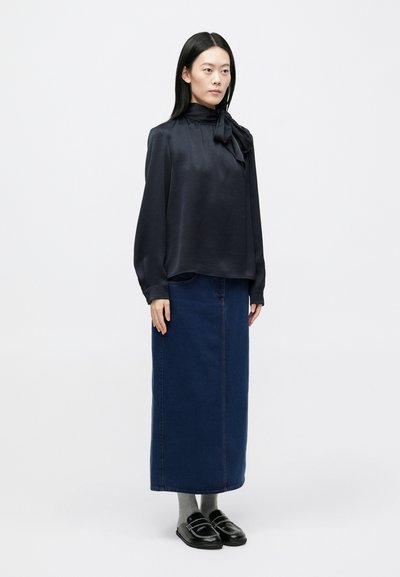 Blusa in raso blu navy con fiocco, abbinata a una lunga gonna di jeans. L'outfit è completato da calzini grigi e scarpe slip-on nere.