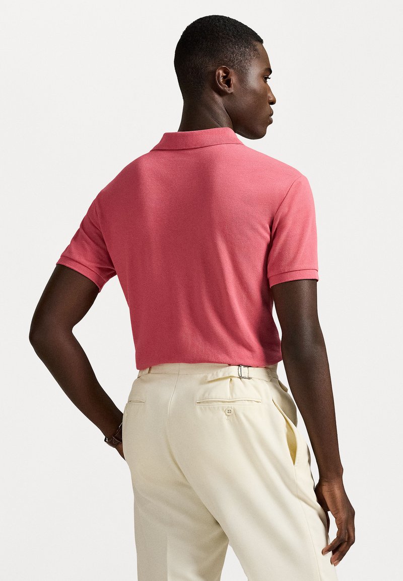 Polo in corallo rosa, maniche corte, tessuto texturizzato. Abbinato a pantaloni beige con tasche posteriori e aperture laterali.