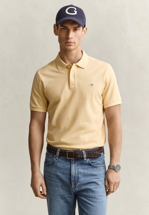 Junger Mann trägt ein beiges Poloshirt, blaue Jeans mit einem braunen geflochtenen Gürtel, eine marineblaue Kappe mit weißem G und eine silberne Armbanduhr.