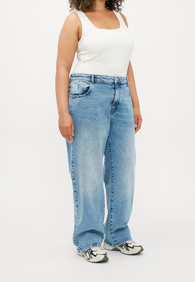 Femme portant un débardeur blanc, un jean bleu clair ample et des chaussures de sport blanches et noires, debout devant un fond uni.