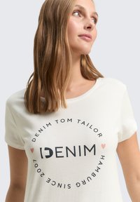 Kreemivärvi t-särk lühikeste varrukatega, millel on ümmargune graafiline print musta ja roosa tekstiga, sealhulgas sõnad "DENIM" ja "Hamburg Since 2007."